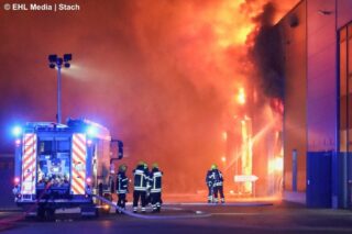 Mehrere Feuerwehrleute in Schutzausrüstung und mit Atemschutz stehen neben einem Löschfahrzeug vor einer brennenden Industriehalle. Aus der Halle schlagen Flammen, und dichter Rauch steigt auf.